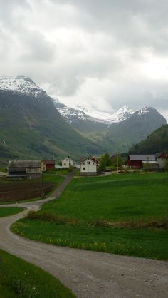 a dirt road in the middle of a green field with snow covered mountains behind it