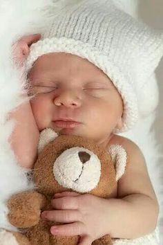 a baby sleeping next to a brown teddy bear wearing a white knitted hat and holding it's head