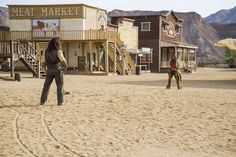 two people are playing frisbee in the sand near some buildings and mountain tops