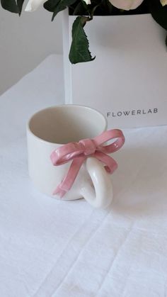 a white cup with a pink bow on it sitting next to a box and flowers