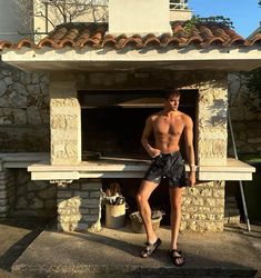 a shirtless man standing in front of an outdoor oven with his hands on his hips