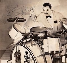 a man in a suit and bow tie sitting on top of a drum kit with his hands up