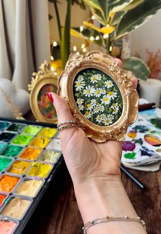 a person holding up a small gold flower brooch in front of paint palettes