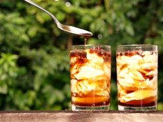 two glasses filled with liquid and ice sitting on top of a table next to a spoon