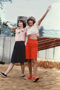 two young women standing next to each other in front of a fence and hay bales