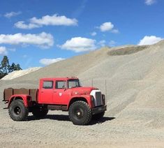 an old red truck parked in the middle of a field with large mounds of sand behind it