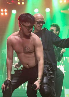 two shirtless men are performing on stage with green lights behind them and one man is holding his arm out