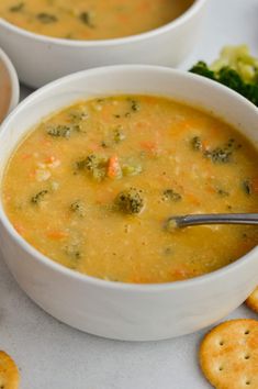 two white bowls filled with soup next to crackers and broccoli on a table