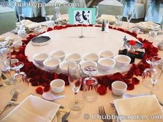 a round table with white dishes and red rose petals on the center, surrounded by silverware