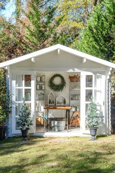 a small white shed with a wreath on the door and shelves full of potted plants