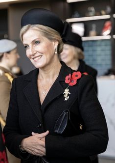 a woman in a black coat and hat standing next to other women wearing red flowers