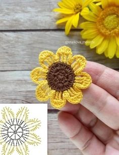 a hand holding a crochet sunflower brooch in front of two yellow flowers