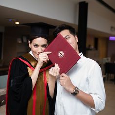 a man and woman in graduation gowns posing for the camera with a red book