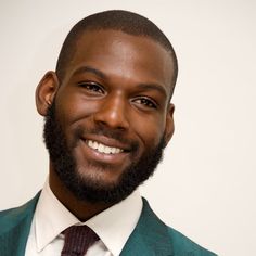 a man in a suit and tie smiling at the camera with a white wall behind him