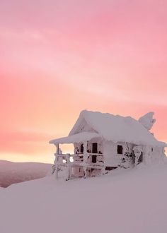 a house covered in snow on top of a snowy hill under a cloudy pink sky