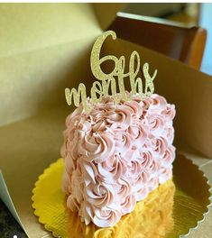 a cake with pink frosting and gold lettering on top sits in a box next to a pair of scissors