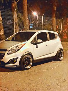 a small white car parked in front of a chain link fence at night with its lights on