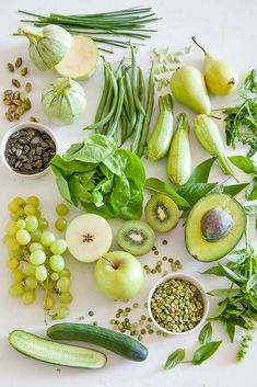 an assortment of fruits and vegetables on a white surface with green leaves, beans, cucumbers, peas, avocados