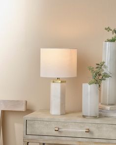 a white table lamp sitting on top of a wooden dresser next to a vase filled with flowers