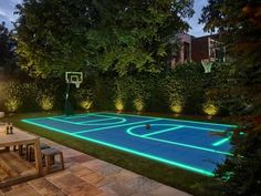 an outdoor basketball court is lit up with blue lights and surrounded by lush greenery