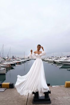 a woman in a long white dress standing on a dock next to some docked boats