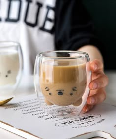 a person holding a coffee cup with a cat face on it's side, while sitting at a table