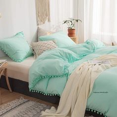 a bed with green sheets and pillows in a room next to a table, chair and potted plant