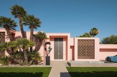 two people walking past a pink house with palm trees in the front yard and a blue car parked on the driveway