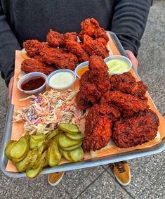 a person holding a tray full of fried chicken and pickles with dipping sauces