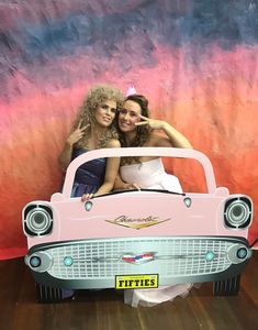 two women pose in front of a pink car