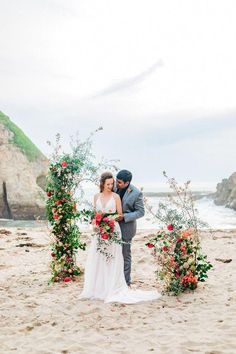 a bride and groom standing on the beach under an arch decorated with flowers for their wedding ceremony