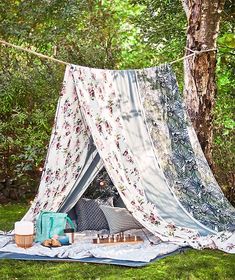 a tent set up in the grass with pillows and blankets on it, next to a tree