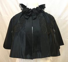 "1890s, 42\" across shoulders, short black satin cape, with black lace applique across cape, with round jet buttons in center of applique.   Hand made black lace frill around neck.  Cape is fastened in front with black silk bow.   Measurements: 42\" across chest.  Width at bottom 112\" Collar to hem 16\" Neck 13 1/2\" Condition excellent" Formal Black Capelet, Black Capelet For Costume, Black Fitted Cape For Formal Occasions, Formal Black Cape With Cape Sleeves, Fitted Black Formal Cape, Elegant Black Cape For Costume, Formal Fitted Black Cape, Black Evening Cape, Black Fitted Capelet