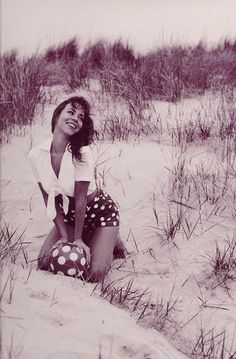 a woman sitting on top of a sandy beach next to grass and sand dunes with trees in the background