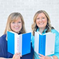 two women holding blue and white books in front of a brick wall