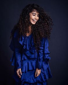 a woman with long curly hair wearing a blue dress and smiling at the camera while standing in front of a dark background