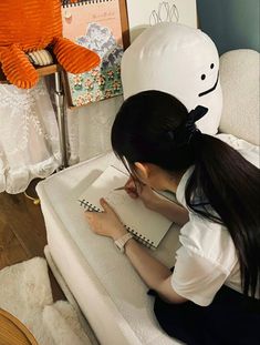 a woman sitting on a couch writing in a notebook with a stuffed animal behind her