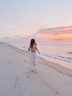 a woman walking on the beach at sunset with her back to the camera, wearing white pants