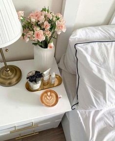 a white nightstand topped with pink flowers next to a night stand and cup of cappuccino