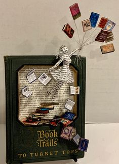 an altered book with books falling off the pages and on top of it is a skeleton holding a bunch of cards