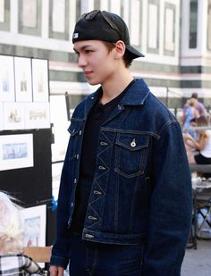 a young man wearing a denim jacket and hat standing in front of an art display