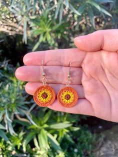 a hand holding a pair of orange and yellow earrings in front of some green plants