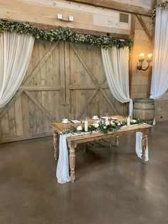 a wooden table with candles and flowers on it in front of a barn door decorated with greenery