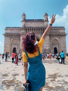 a woman in overalls is waving to the sky at an old building with many people around her