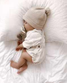 a baby laying on top of a bed next to a teddy bear wearing a hat