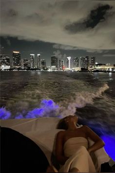 a woman laying on top of a boat in the ocean next to a city at night