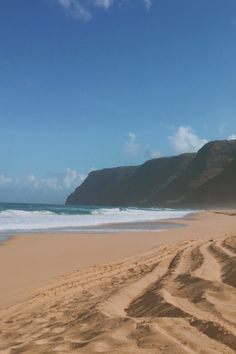 a sandy beach next to the ocean under a blue sky with clouds and mountains in the background