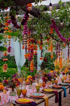 an outdoor dining area decorated with flowers and hanging candles, plates and glasses on the table