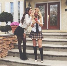 two young women standing on the steps in front of a house looking at their cell phones