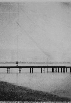 black and white photograph of person walking on pier over water with fog in the background